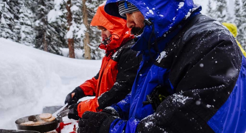 two veterans wearing snow gear cook food at their snowy campsite